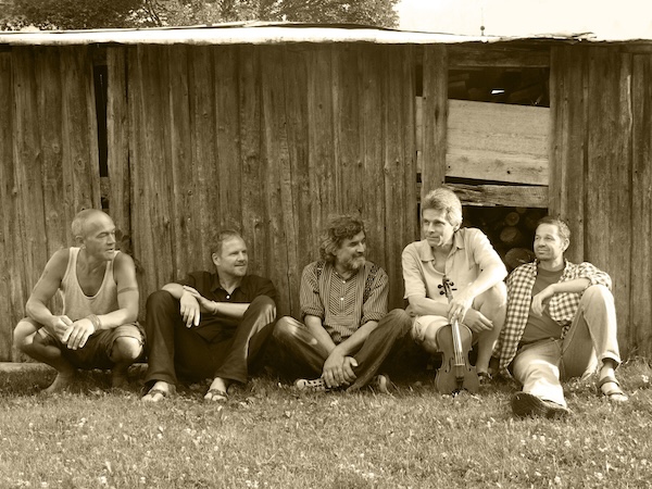 Fünf Männer sitzen entspannt vor einer alten Holzwand auf einer Wiese
