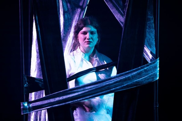 Schauspielerin in weißem Hemd hinter dunklen Stoffbahnen auf einer Bühne